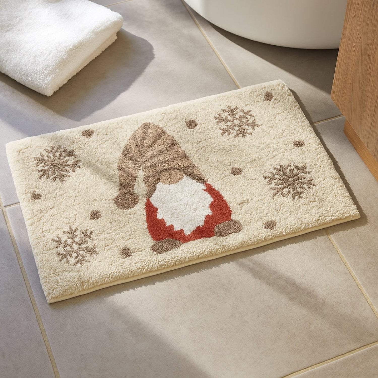 A rectangular beige bath mat with a festive gnome design and snowflakes, placed on a tiled bathroom floor next to a folded white towel.