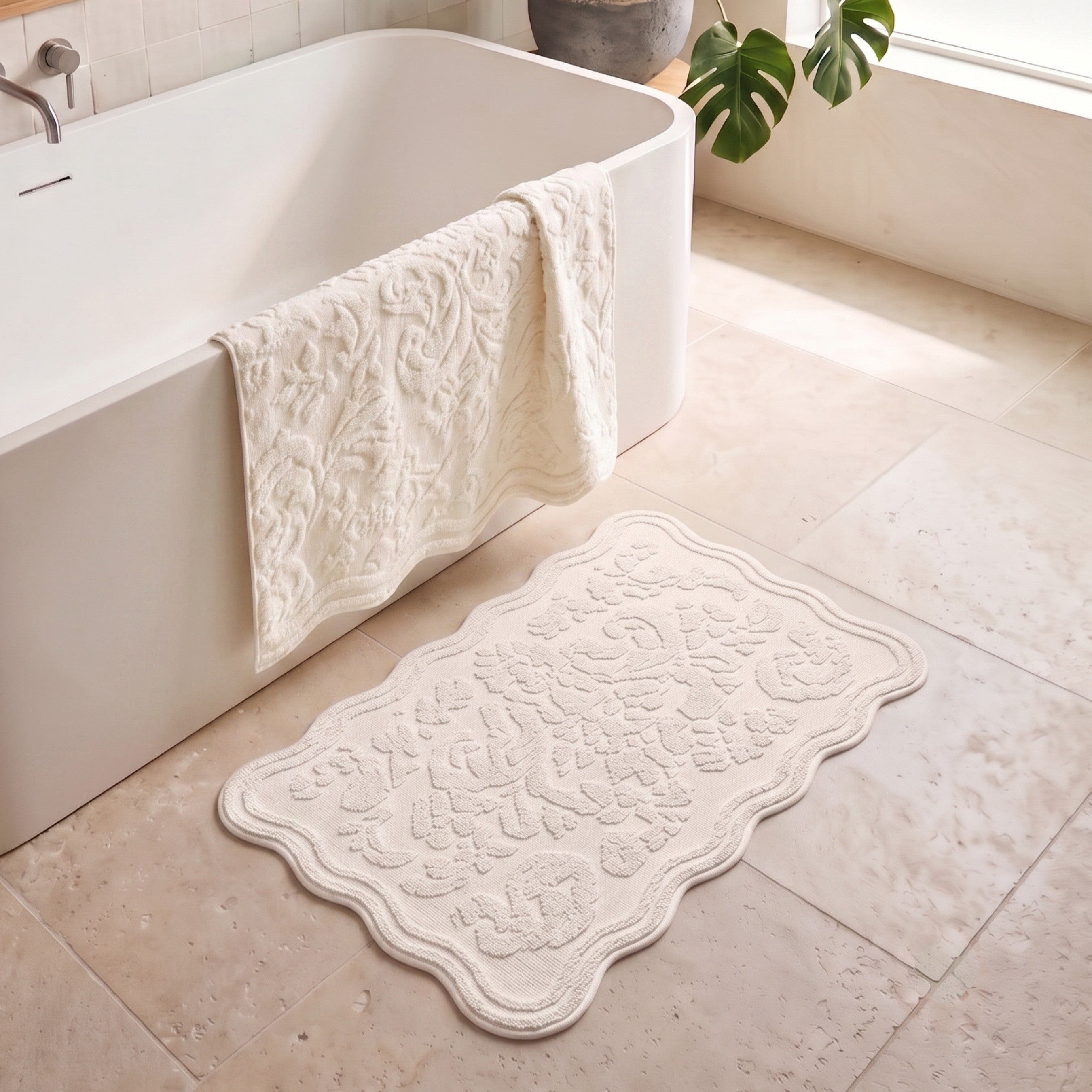A cream scalloped bath mat with a raised floral damask pattern lies on a beige tiled bathroom floor next to a white bathtub with a matching bath towel draped over the edge.