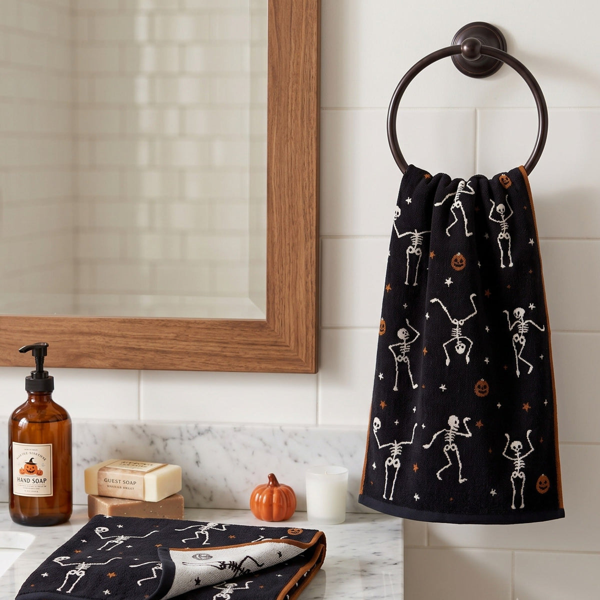 A black Halloween-themed towel with white skeleton and orange pumpkin prints hangs on a dark metal ring hook on a white tiled bathroom wall. A wooden-framed mirror, soap dish, pump bottle, and small decorative pumpkins are visible on the counter.