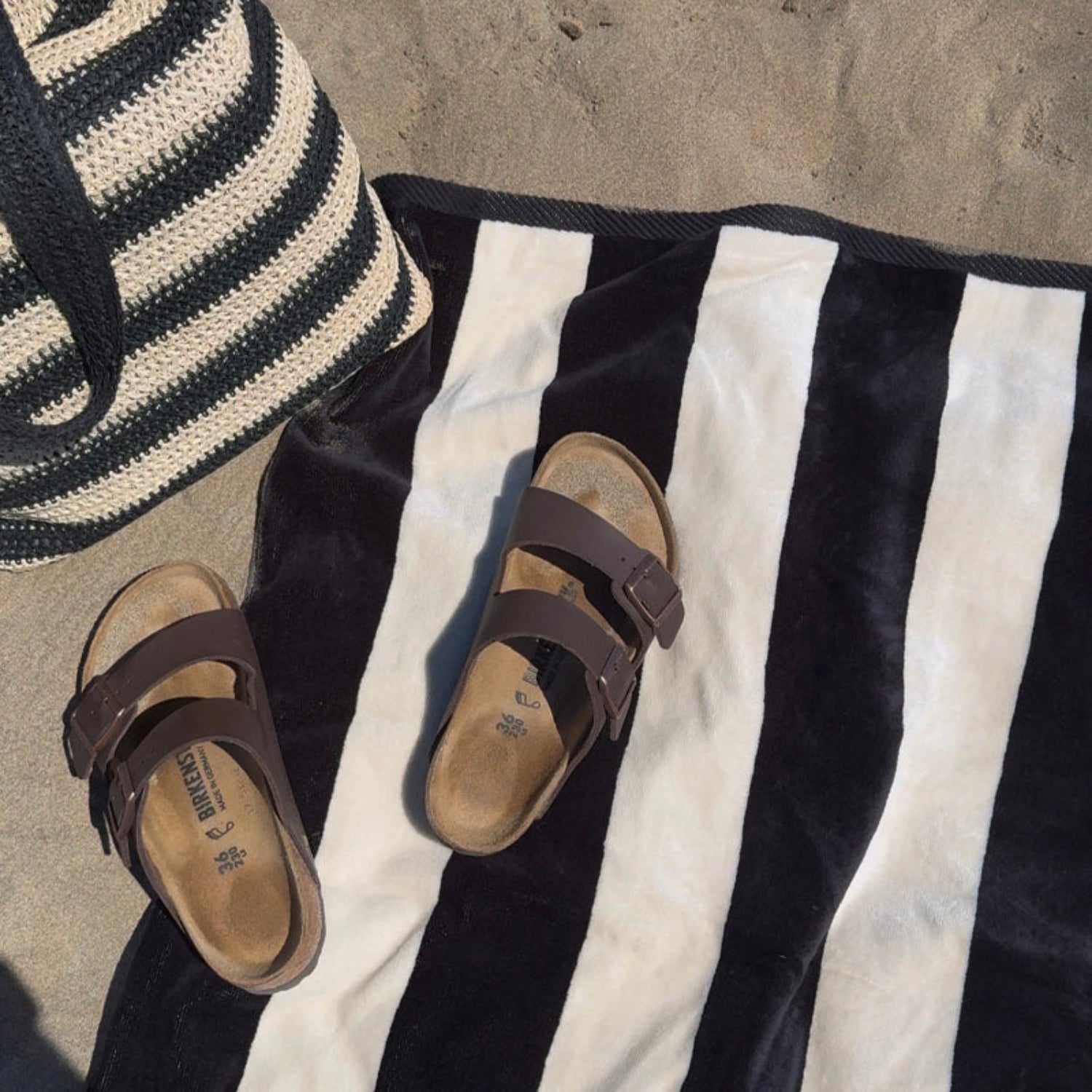 Brown sandals on a black and beige striped towel with a woven bag in the background.