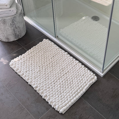White textured bath mat on a bathroom floor with a shower in the background