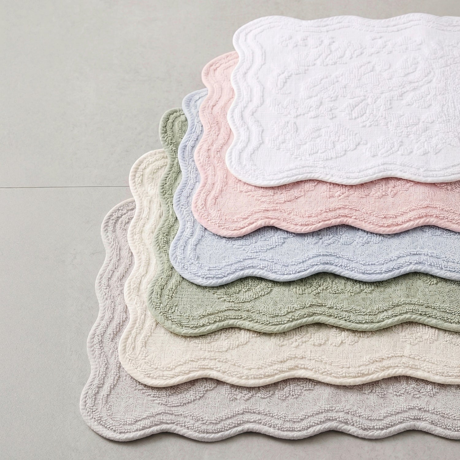Six scalloped bath mats in soft pastel colours stacked on a light grey floor. The colours visible from top to bottom are white, pink, light blue, sage green, cream, and light grey.