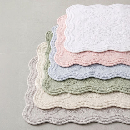 Six scalloped bath mats in soft pastel colours stacked on a light grey floor. The colours visible from top to bottom are white, pink, light blue, sage green, cream, and light grey.