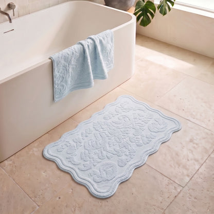A light blue scalloped bath mat with an embossed floral pattern lies on a beige tiled bathroom floor next to a white bathtub, with a matching light blue towel draped over the tub edge.