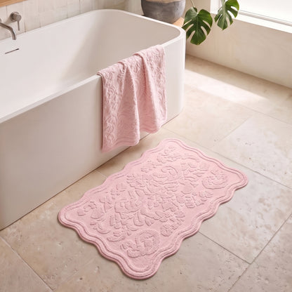 A pink scalloped bath mat with an ornate embossed floral pattern lies on a beige tiled bathroom floor next to a white bathtub, with a matching pink towel draped over the tub edge.