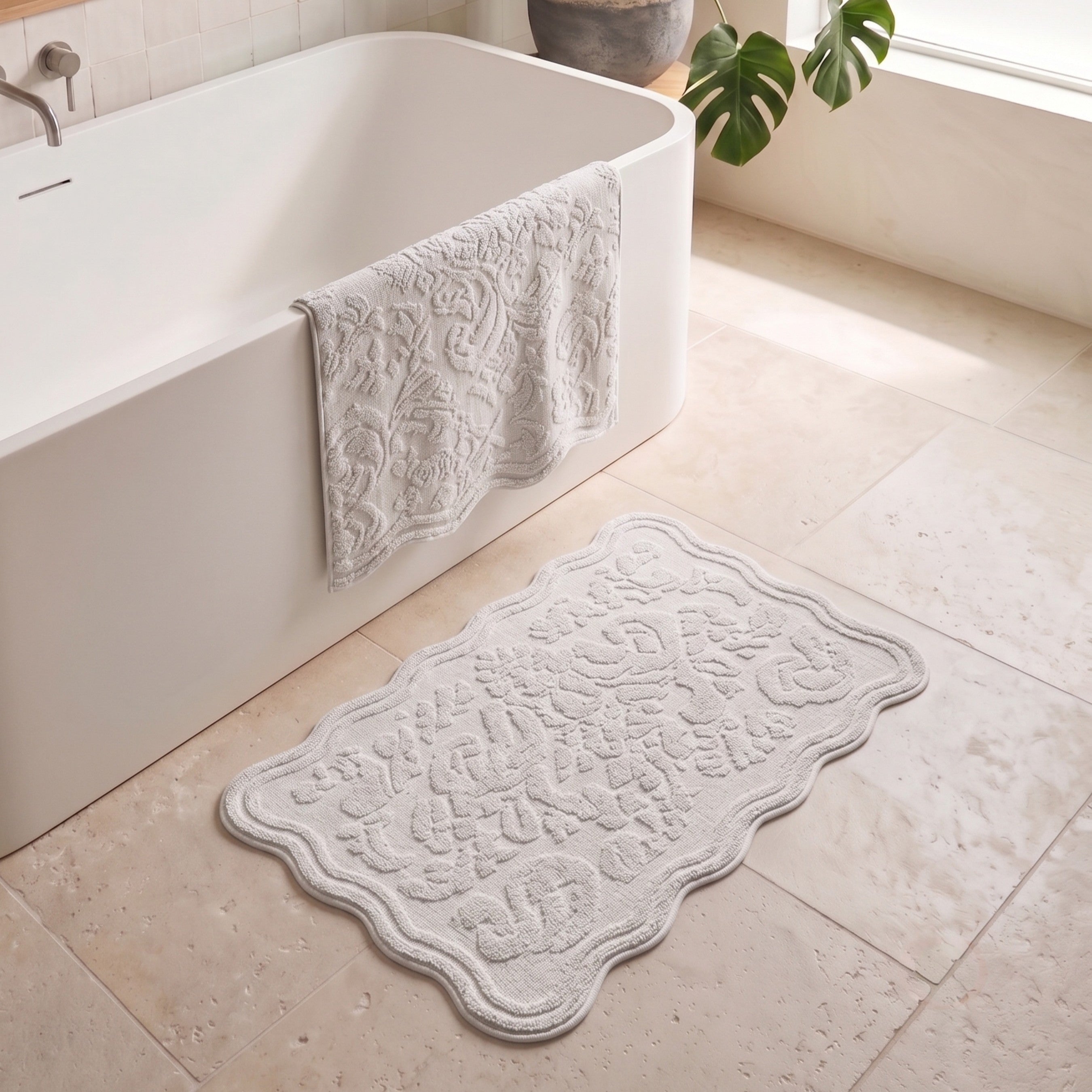 A rectangular bath mat with a scalloped edge and raised floral pattern in a light grey color, placed on a beige tiled bathroom floor next to a white freestanding bathtub with a matching grey towel draped over the tub.
