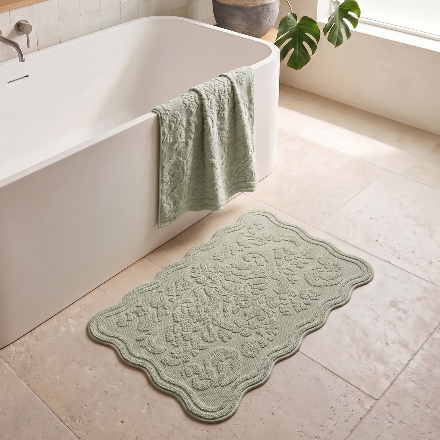 A light sage green scalloped bath mat with an embossed floral pattern lies on a beige tiled bathroom floor next to a white freestanding bathtub, with a matching towel draped over the tub edge.
