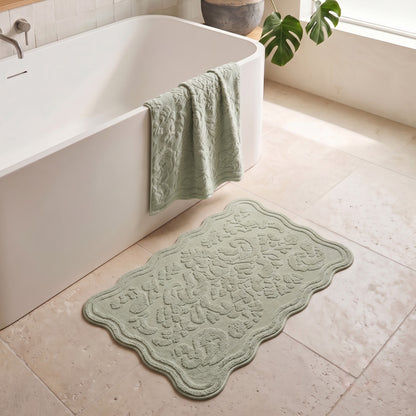 A light sage green scalloped bath mat with an embossed floral pattern lies on a beige tiled bathroom floor next to a white freestanding bathtub, with a matching towel draped over the tub edge.