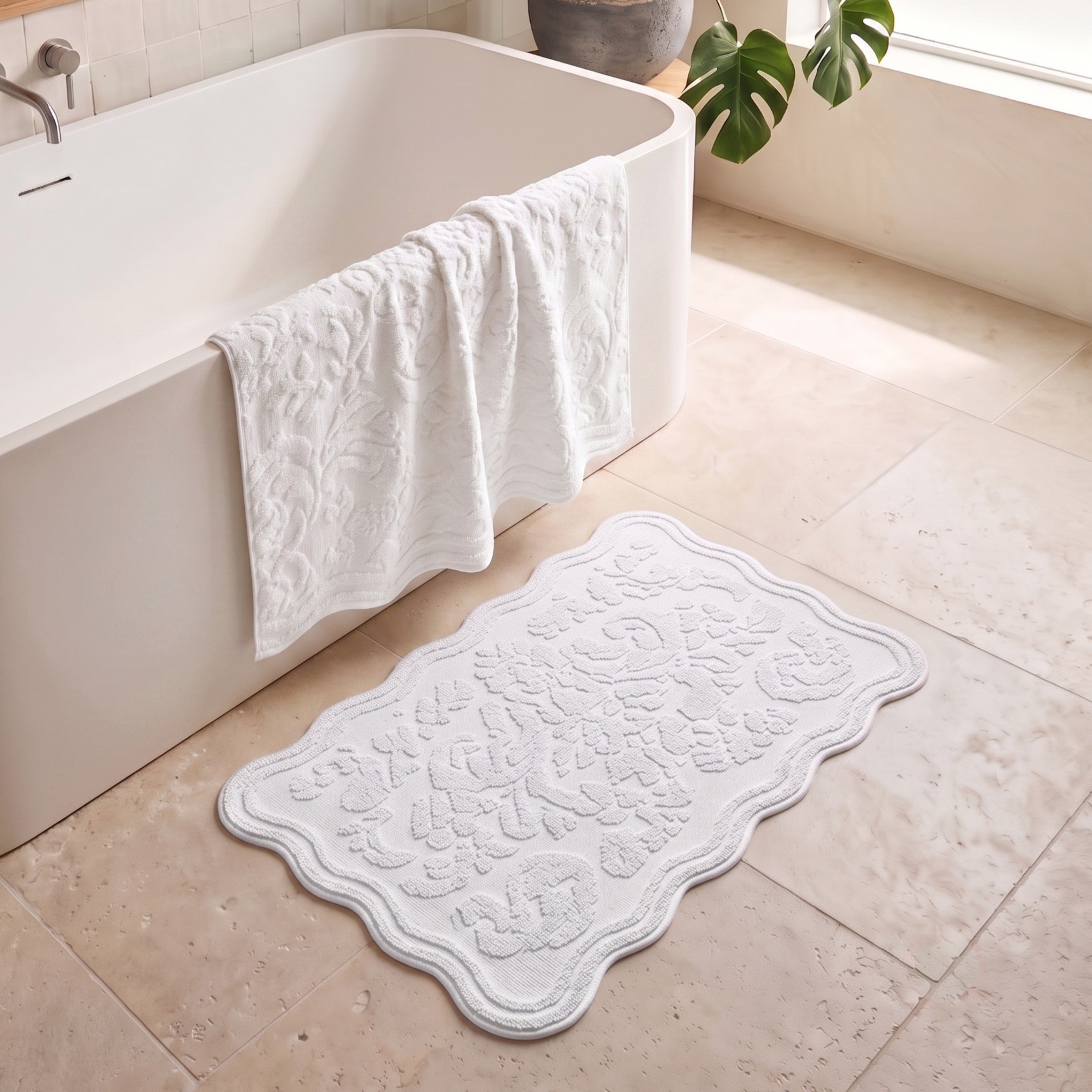 A white textured bath mat with a scalloped edge and embossed floral pattern lies on a beige tiled bathroom floor next to a white freestanding bathtub, with a white towel draped over the tub and a green plant leaf in the background.