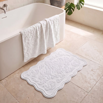 A white textured bath mat with a scalloped edge and embossed floral pattern lies on a beige tiled bathroom floor next to a white freestanding bathtub, with a white towel draped over the tub and a green plant leaf in the background.