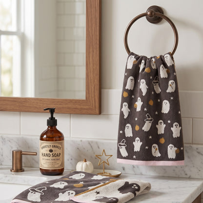 A Halloween-themed hand towel hanging on a brass ring holder on a bathroom wall, with a matching folded towel on the counter below. The towels are dark brown with white ghosts, gold moons and stars, and pink trim.