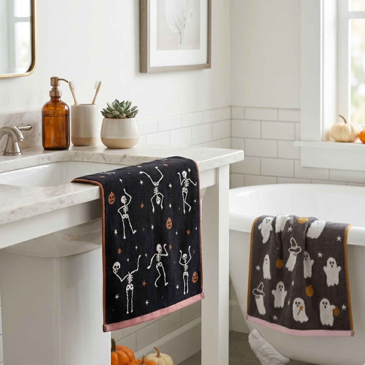 Two Halloween-themed bath towels hanging over a white bathroom vanity and tub. One towel is black with white skeleton figures and small orange pumpkin accents, finished with a pink border.