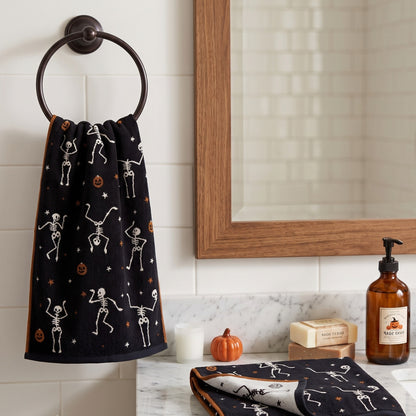 A black Halloween-themed towel with white skeleton and orange pumpkin prints hangs on a dark metal ring hook on a white tiled bathroom wall. A wooden-framed mirror, soap dish, pump bottle, and small decorative pumpkins are visible on the counter.