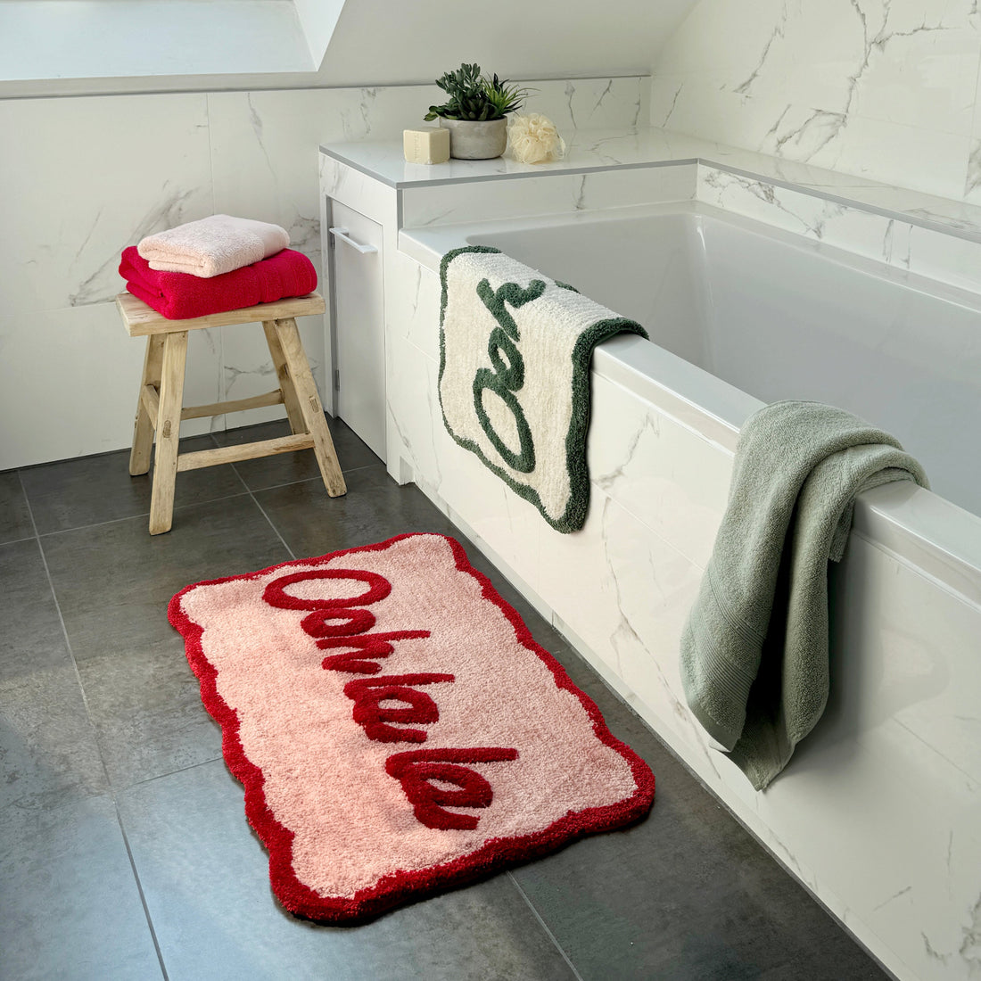 Ooh la la slogan scalloped bath mats next to a bath tub, hand tufted in pink/red and cream/green.