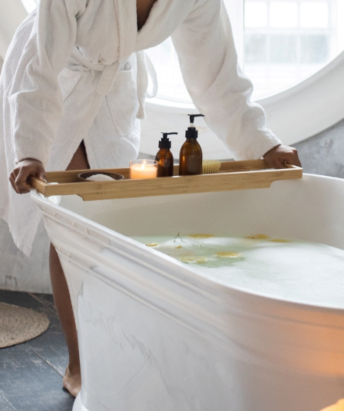 Person in a bathrobe holding a bamboo tray with bath products over a bathtub filled with water.