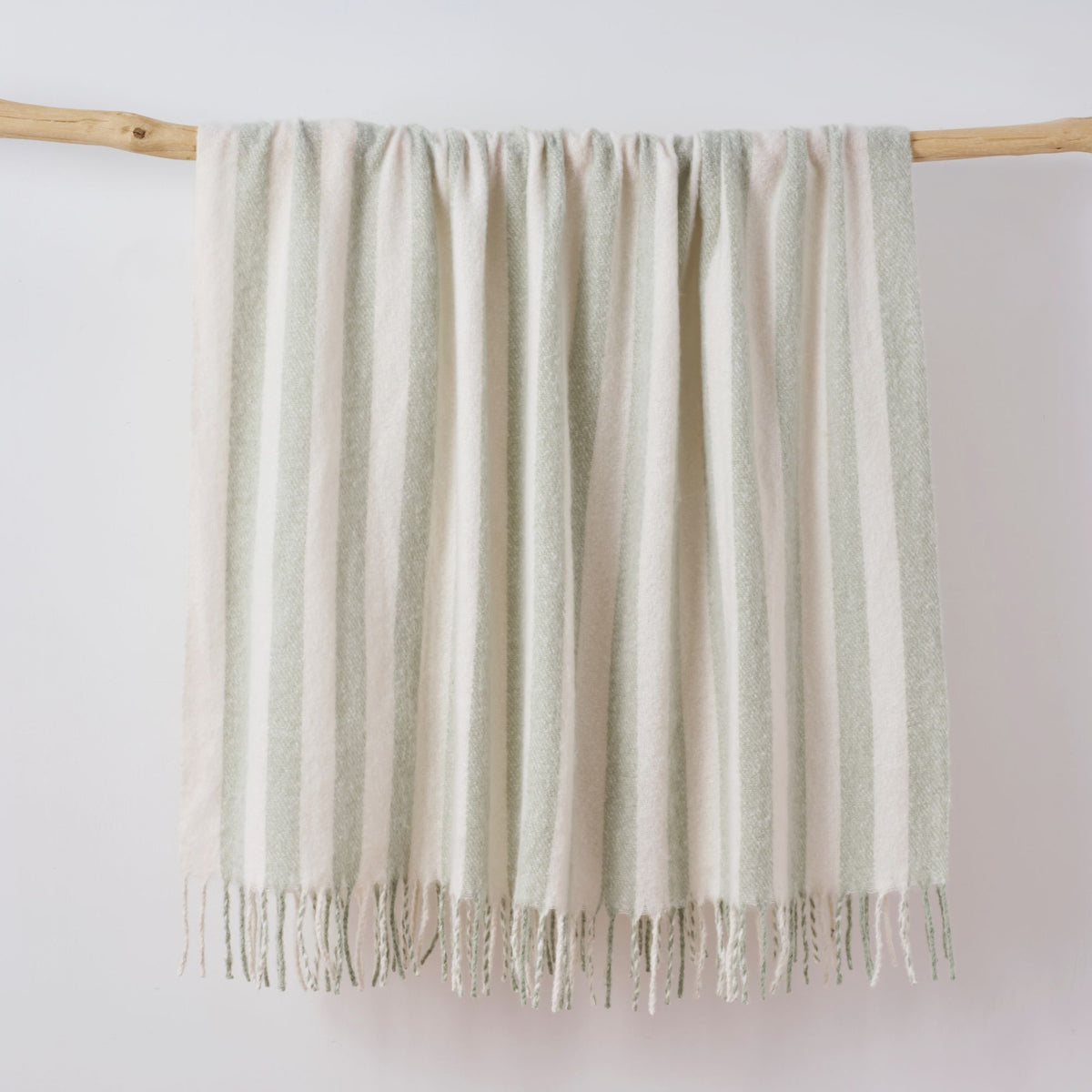 A striped faux mohair throw blanket in sage green and off white hangs from a light wood dowel against a plain white wall.