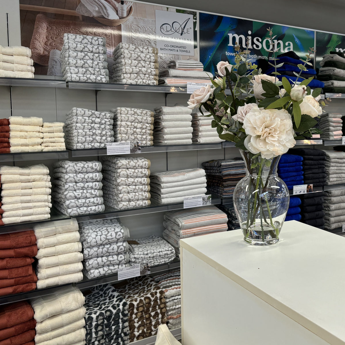 A styled retail interior showing shelves stocked with folded textiles and bedding in neutral tones, with a clear glass vase of pale cream flowers on a white counter in the foreground. A large sign in the background reads "misona.