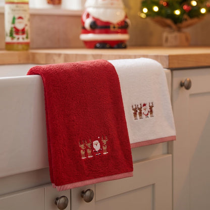 Two Christmas-themed towels draped over a white farmhouse sink in a kitchen setting. One towel is bright red with embroidered Santa Claus and reindeer, and the other is white with matching embroidery and a red striped border.