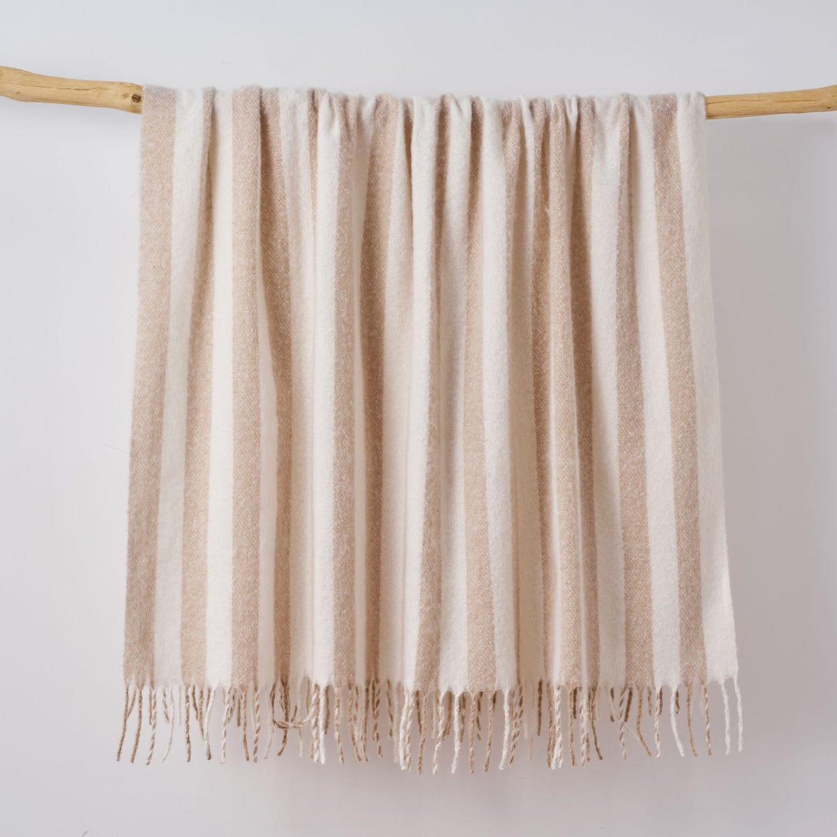 A cream and beige striped faux mohair blanket with fringe tassels hanging from a light wooden dowel against a plain white wall.