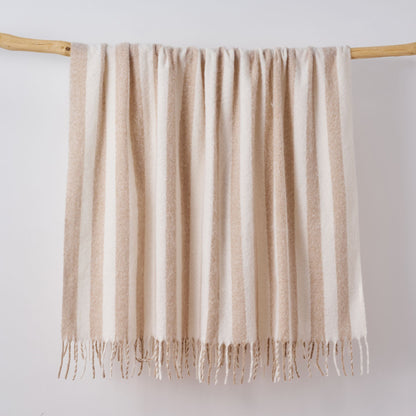 A cream and beige striped faux mohair blanket with fringe tassels hanging from a light wooden dowel against a plain white wall.