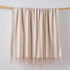 A cream and beige striped faux mohair blanket with fringe tassels hanging from a light wooden dowel against a plain white wall.